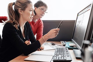 Two women are looking at a computer screen where one woman is pointing at the screen with a black pencil