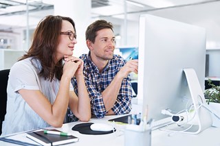 A man and a woman are sitting in front of a computer screen where the man is pointing at the screen with a black pencil